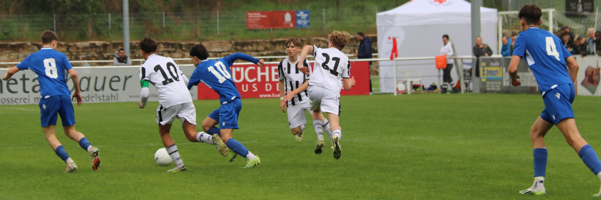 Eine Spielsituation in einem Fußballspiel. Zwei Juniorenmannschaften, eine in blau, eine in weiß, spielen gegeneinander. In der Bildmitte kämpfen ein Spieler in blau und ein Spieler in weiß um den Ball.