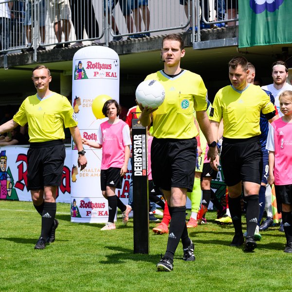 Schiedsrichtergespann, das beim Pokalfinale aus dem Tunnel läuft. Der Schiri in der Mitte hat den Ball in der Hand, hinter ihm laufen die Spieler der beiden Mannschaften mit Einlaufkindern ein.