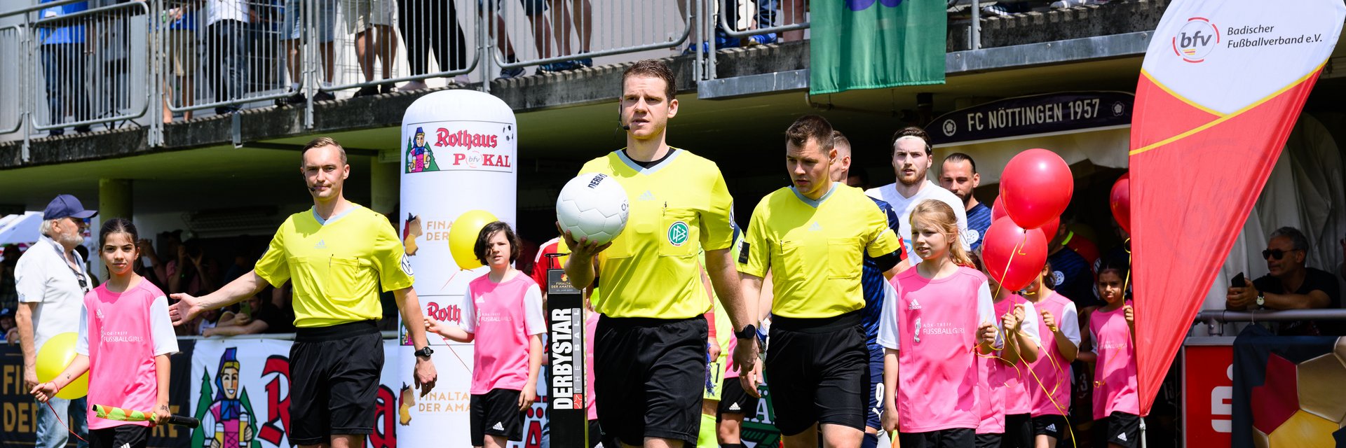 Schiedsrichtergespann, das beim Pokalfinale aus dem Tunnel läuft. Der Schiri in der Mitte hat den Ball in der Hand, hinter ihm laufen die Spieler der beiden Mannschaften mit Einlaufkindern ein.