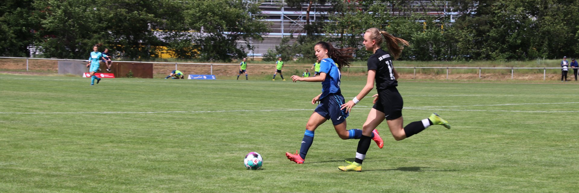 Zwei Spielerinnen, die einen Zweikampf führen. Eine von ihnen trägt ein schwarzes Trikot, die andere ein Blaues.