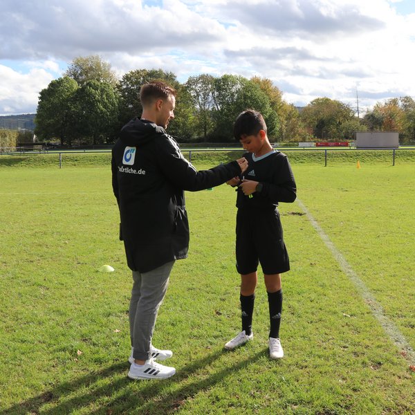 Erwachsener und einen jugendlicher Schiedsrichter. Sie stehen gemeinsam auf einem Fußballplatz, der ältere Schiri hilft dem jüngeren, seine Karten in der Brusttasche zu verstauen.