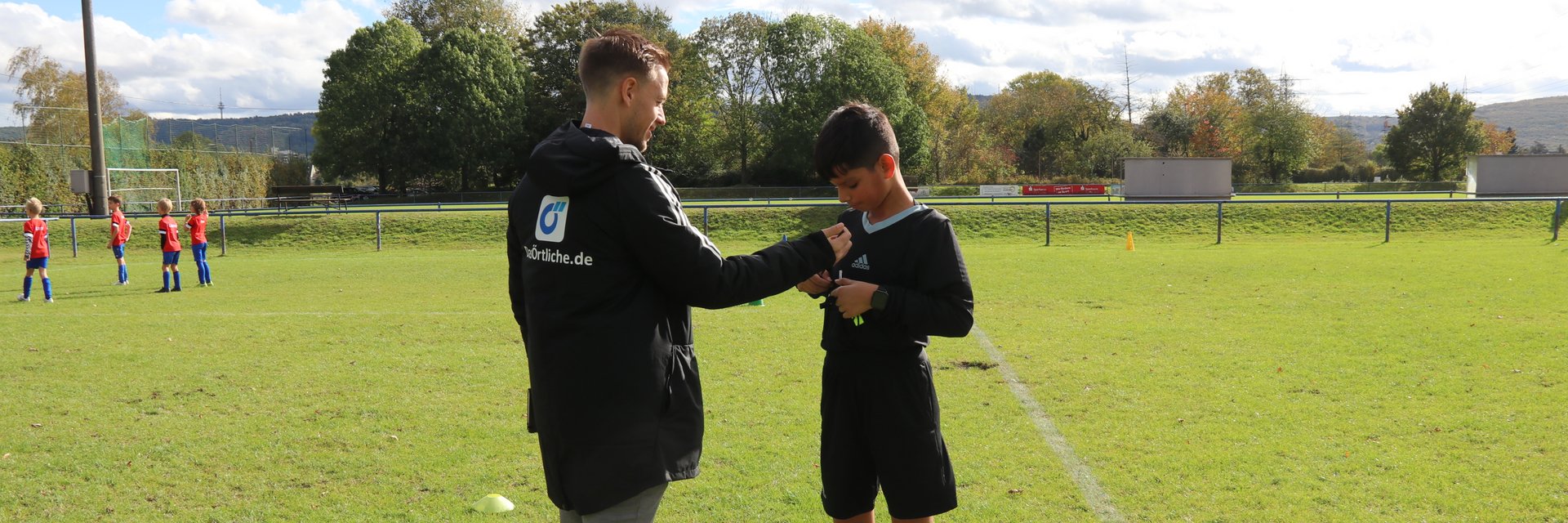 Erwachsener und einen jugendlicher Schiedsrichter. Sie stehen gemeinsam auf einem Fußballplatz, der ältere Schiri hilft dem jüngeren, seine Karten in der Brusttasche zu verstauen.