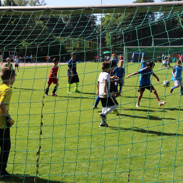 Kinder, die Fußball spielen. Ein Kind schießt, der Torwart steht bereit. Es ist von hinten durch das Tornetz fotografiert.