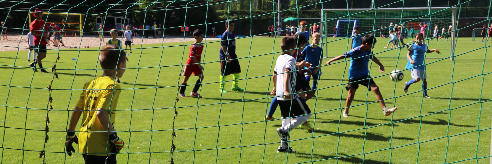 Kinder, die Fußball spielen. Ein Kind schießt, der Torwart steht bereit. Es ist von hinten durch das Tornetz fotografiert.