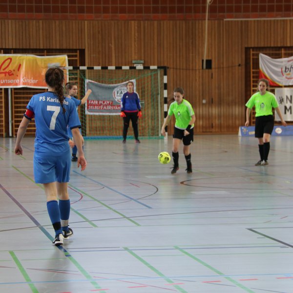 Vier Spielerinnen, die in einer Halle Futsal spielen. Zwei von ihnen haben ein blaues Trikot an, die anderen beiden ein grünes.
