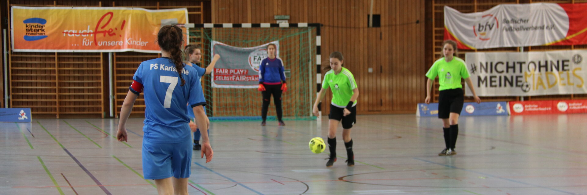Vier Spielerinnen, die in einer Halle Futsal spielen. Zwei von ihnen haben ein blaues Trikot an, die anderen beiden ein grünes.