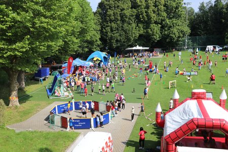 Zahlreiche Kinder, die auf dem Rasenplatz der Sportschule Schöneck an verschiedenen Aktivitäten, wie z.B. einer Hüpfburg Spaß haben.