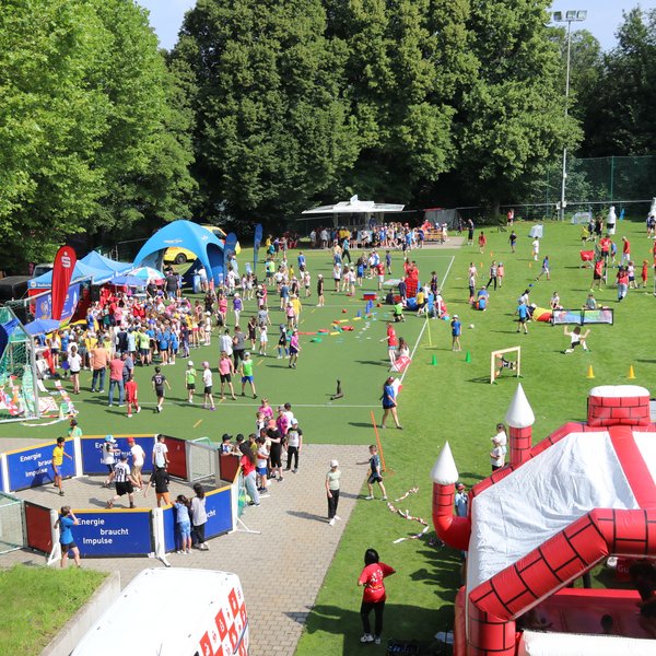 Zahlreiche Kinder, die auf dem Rasenplatz der Sportschule Schöneck an verschiedenen Aktivitäten, wie z.B. einer Hüpfburg Spaß haben.