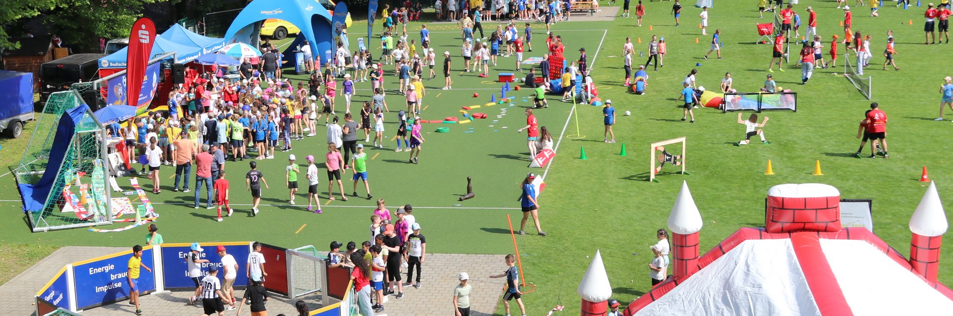 Zahlreiche Kinder, die auf dem Rasenplatz der Sportschule Schöneck an verschiedenen Aktivitäten, wie z.B. einer Hüpfburg Spaß haben.