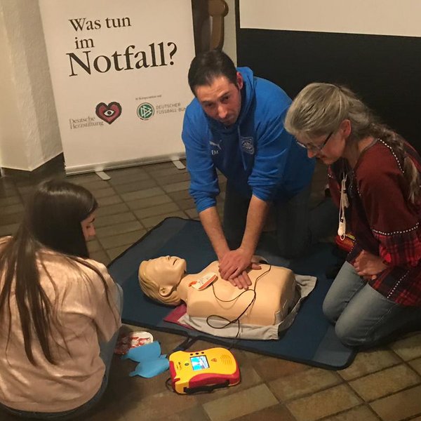 Drei Personen, die auf dem Boden sitzen. Der Mann in der Mitte gibt einer Puppe eine Herzdruckmassage, die beiden Frauen neben ihm beobachten ihn dabei.