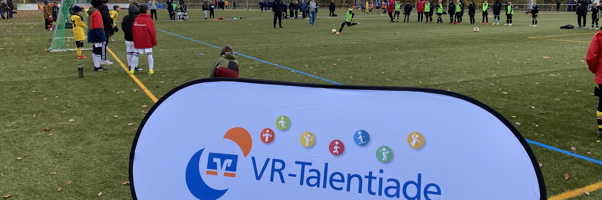 Banner der VR-Talentiade, im Hintergrund stehen Kinder Schlange um aufs Tor zu schießen.