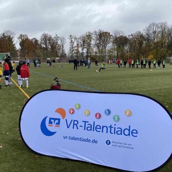 Banner der VR-Talentiade, im Hintergrund stehen Kinder Schlange um aufs Tor zu schießen.