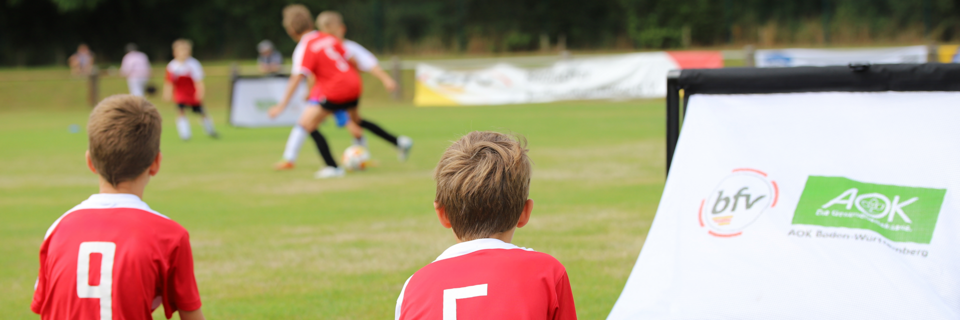 zwei Jungen in roten Trikots, die auf dem Rasen sitzen und andere Kinder beim Fußballspielen beobachten.