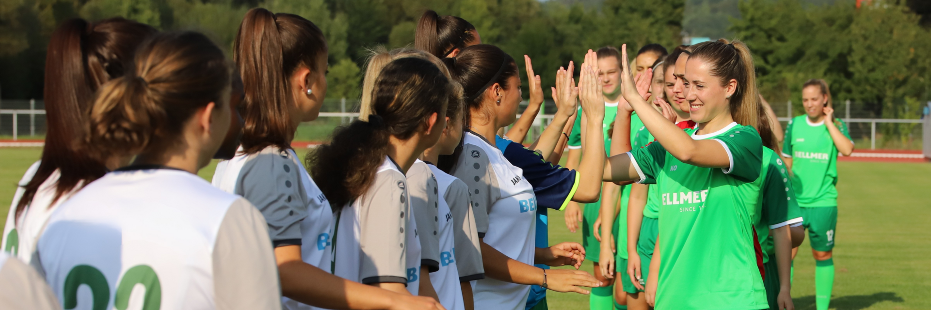 Zwei Frauenmannschaften, die sich nach dem Einlaufen vor dem Spiel abklatschen. Die Mannschaft die steht, trägt weiße Trikots, die Andere Grüne.