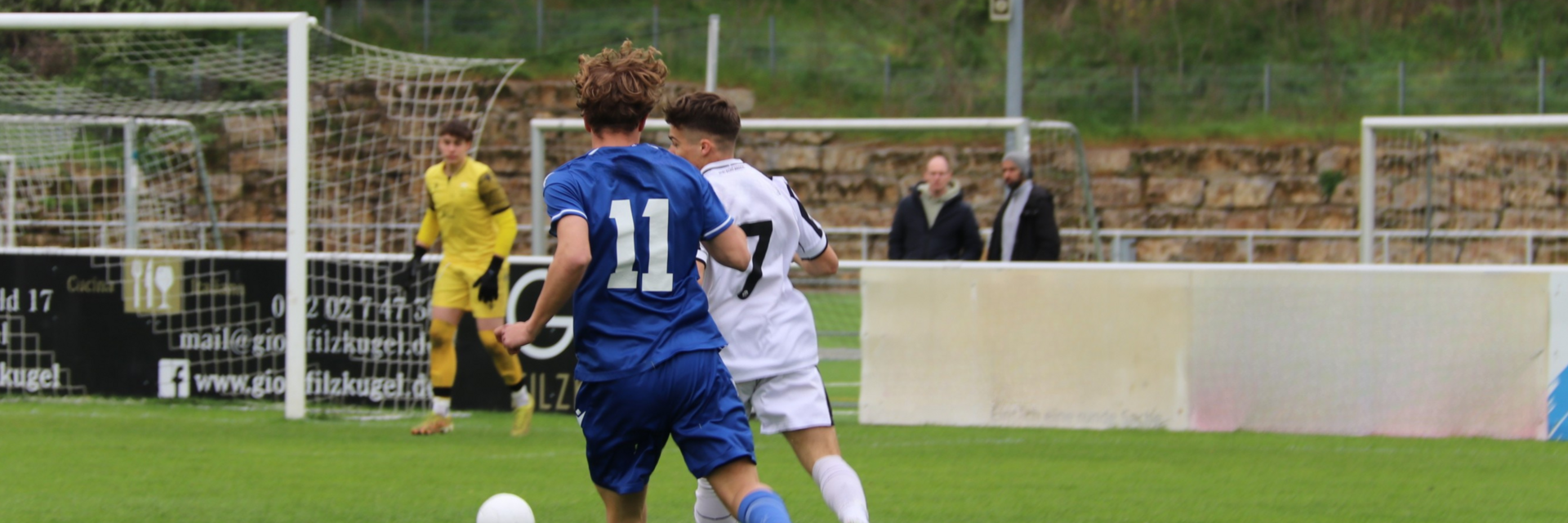 Eine Spielsituation eines Fußballspiels. Ein Spieler in einem weißen Trikot und ein Spieler in einem blauen Trikot kämpfen um den Ball. Im Hintergrund sieht man einen Torhüter in einem gelben Trikot.