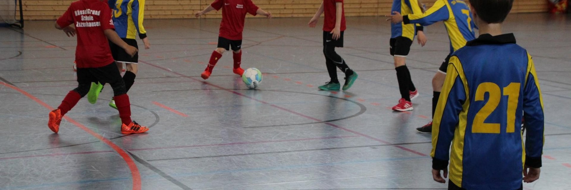 Acht Kinder, die in einer Halle Fußball spielen. In der Bildmitte hat ein Junge in einem roten Trikot den Ball am Fuß. 