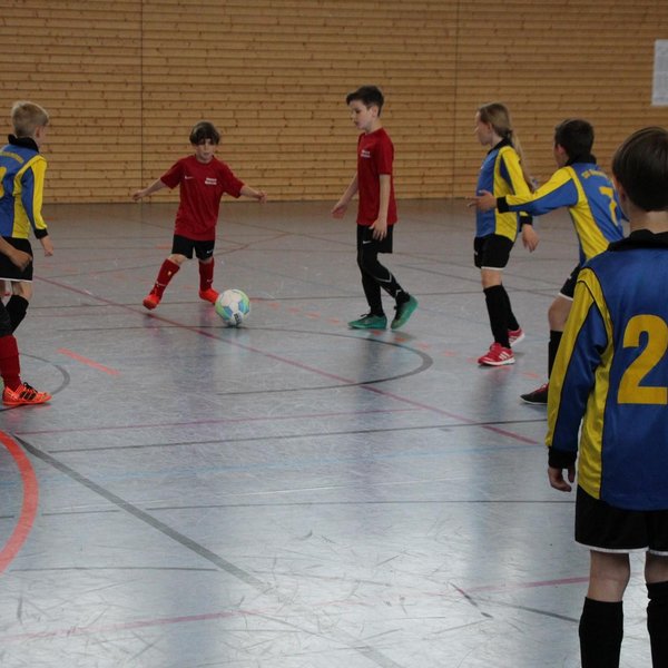 Acht Kinder, die in einer Halle Fußball spielen. In der Bildmitte hat ein Junge in einem roten Trikot den Ball am Fuß. 