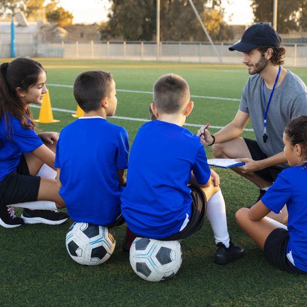 Ein Fußballtrainer, der sich mit vier Kindern, die seine Spieler*innen sind, unterhält. Die Kinder sitzen auf dem Boden oder auf Bällen vor ihm, er sitzt in der Hocke.