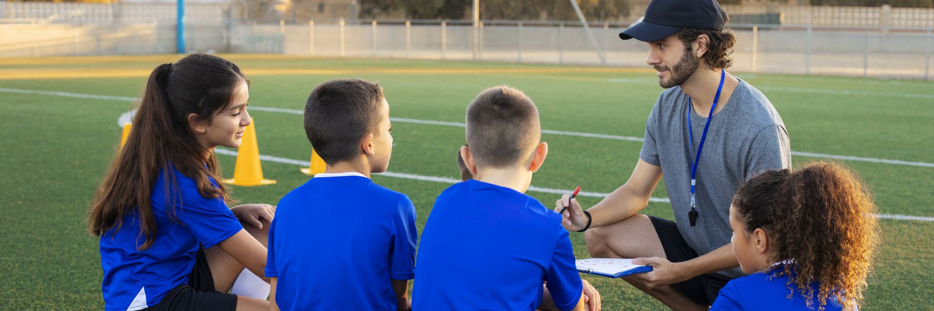 Ein Fußballtrainer, der sich mit vier Kindern, die seine Spieler*innen sind, unterhält. Die Kinder sitzen auf dem Boden oder auf Bällen vor ihm, er sitzt in der Hocke.