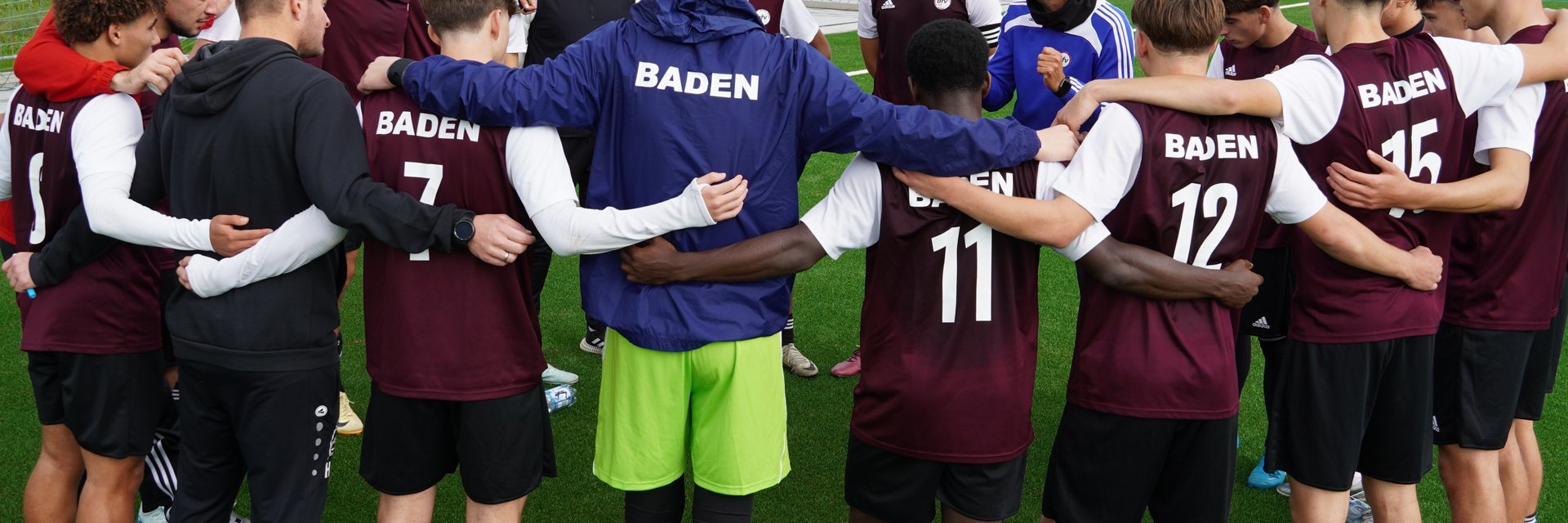 Eine bfv-Juniorenauswahl. Die Jungen stehen in einem Kreis und haben die Arme umeinander gelegt, während der Trainer in der Mitte eine Ansprache hält.