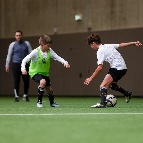 Eine Spielszene aus einem DFB Trainingsdialog. Zwei Spieler führen einen Zweikampf, ein Trainer beobachtet sie im Hintergrund.