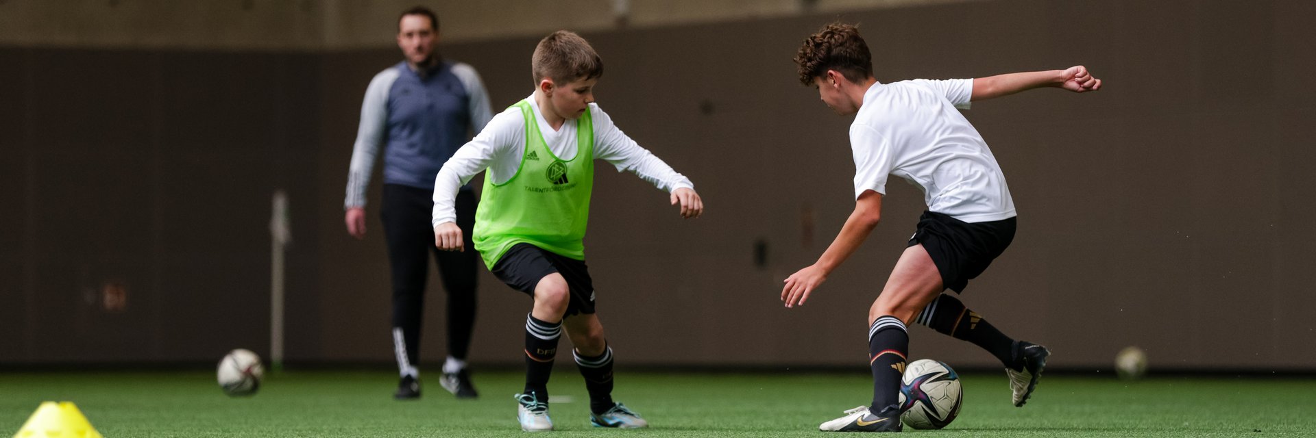 Eine Spielszene aus einem DFB Trainingsdialog. Zwei Spieler führen einen Zweikampf, ein Trainer beobachtet sie im Hintergrund.