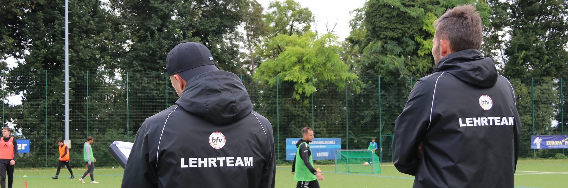 Zwei Mitglieder des bfv-Lehrteams, die am Spielfeldrand stehen und eine Trainerlizenz-Lehrprobe bewerten. Der rechte Trainer beobachtet das Spielgeschehen, während der Linke Notizen schreibt.