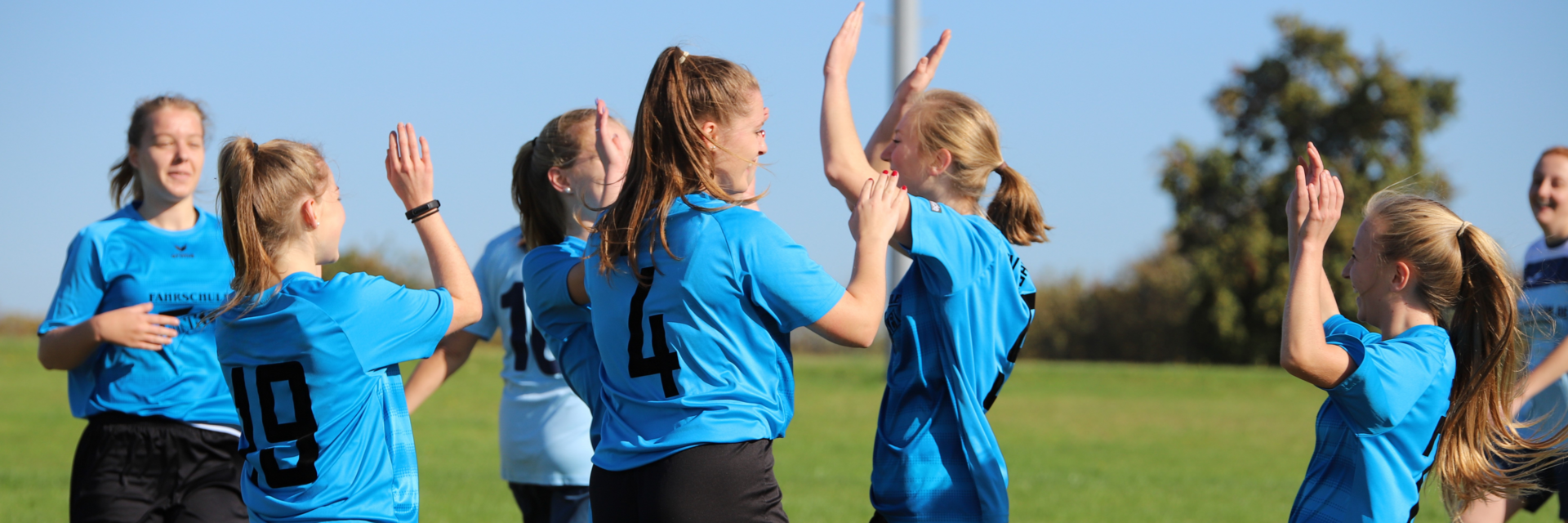 Junge Fußballspielerinnen in blauen Trikots, die jubeln. Sie klatschen sich gegenseitig ab und lächeln.